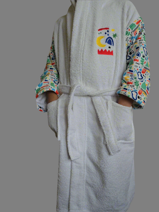 A child wearing a white bathrobe with colorful nature-themed patterns on the sleeves, standing in a bathroom setting.